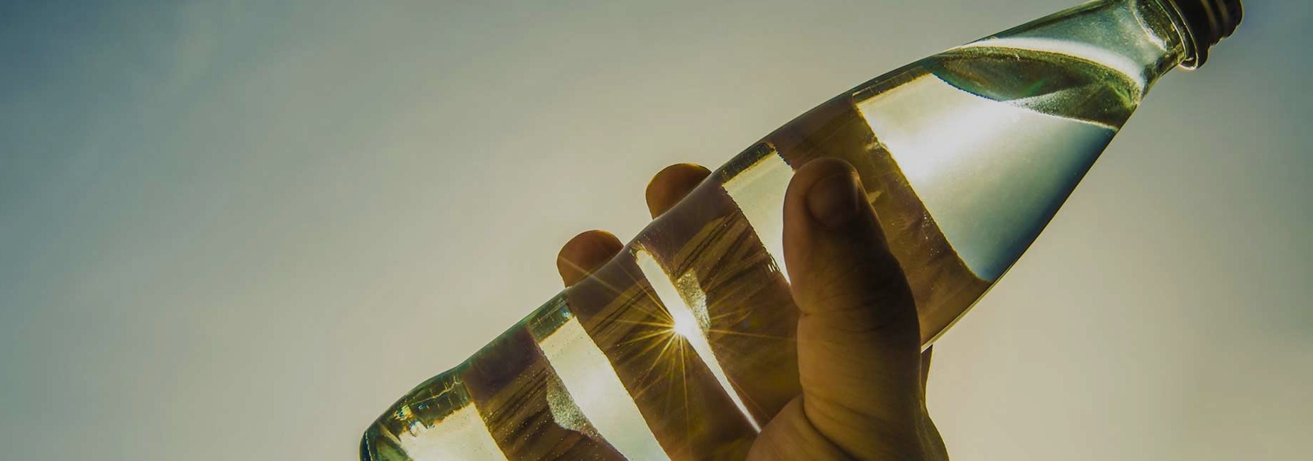 Hand holding glass bottle against blue sky and bright sun, software solutions for manufacturing industry, O-I Glass | DXC Technology Customer Stories